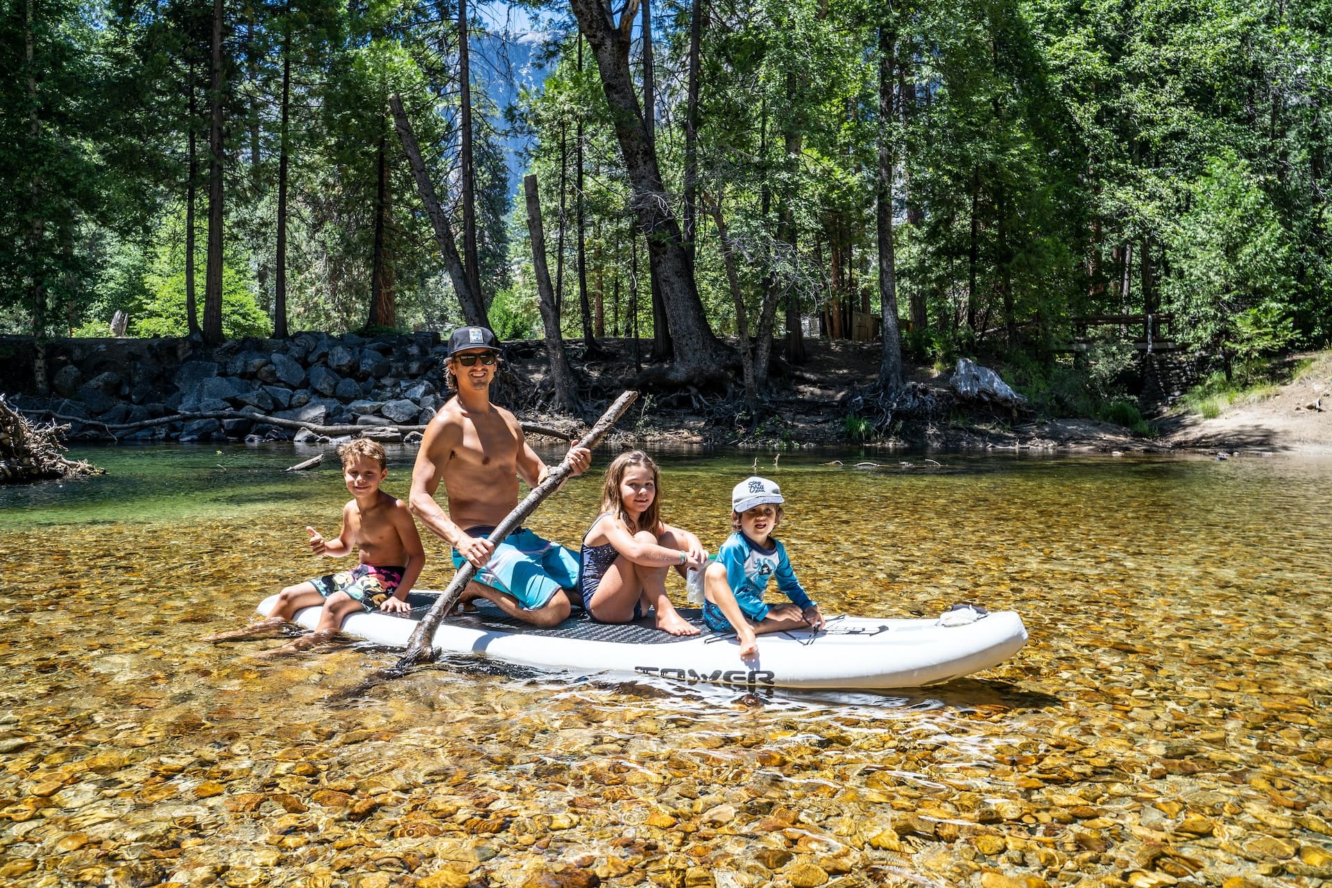 A happy man and three kids playing on a paddle board in the middle of a small lake.